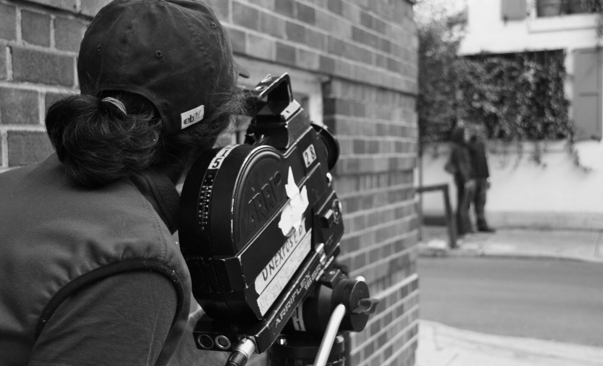 Woman in a baseball cap looking through the eye piece of an Arriflex film camera.